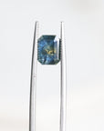 A loose rectangular blue gemstone with internal inclusions, held between silver metal tweezers against a plain white background.