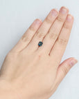 A close-up of a hand wearing a small rectangular dark blue gemstone ring on the ring finger, set against a plain light gray background.