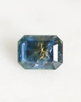 A rectangular emerald-cut blue gemstone with natural inclusions and a gold-colored matrix core, photographed on a clean white background.