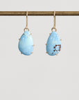 Pair of blue teardrop earrings on a gold earring stand against a light gray background
