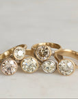 Five gold rings with round clear gemstones, arranged together on a light marble surface.