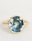 An antique-style ring featuring a large oval blue sapphire with inclusions, surrounded by small white diamonds in a silver setting, with a yellow gold band visible on either side, shot against a plain off-white background.