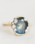 A vintage-style ring featuring a large oval blue gemstone in a gold-toned setting, accented with small white stones, photographed on a light neutral background.