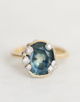 A vintage-style yellow gold ring featuring a large oval blue gemstone surrounded by small clear accent stones, set against a plain light background.