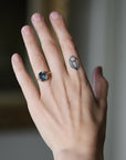 A close-up of a person's hand wearing a gold ring with a dark oval gemstone, alongside a small geometric tattoo on the side of the hand.