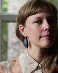 A close-up portrait of a woman with brown hair and bangs wearing a patterned cream blouse and a blue teardrop-shaped dangle earring with a gold-toned setting, posed in front of a soft-focus window.