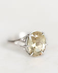 A silver ring featuring an oval-cut, faceted pale yellow gemstone in a prong setting, shown on a soft, light off-white background.