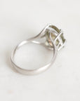A silver ring with a unique interlocking openwork design on the band, holding a small pale green gemstone, shown on a light off-white background.
