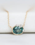 A delicate gold chain necklace with a pendant featuring a faceted teal-blue gemstone and small barnacles, displayed on a soft white background.