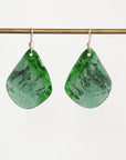 A pair of teardrop-shaped green stone dangle earrings with silver hooks, hanging from a thin horizontal brass rod against a plain light background.