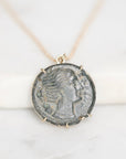 An ancient coin pendant necklace featuring a profile portrait of a woman, set in a gold-toned bezel and suspended from a delicate gold chain against a white marble background.