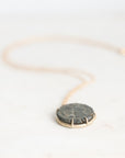 A delicate gold chain necklace with a round, dark gray stone pendant set in a gold bezel, resting on a light off-white marble surface.