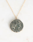 A gold chain necklace featuring a circular pendant made from an ancient, oxidized coin with a profile portrait, displayed on a light marble surface.