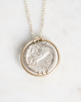A silver pendant necklace featuring a circular coin-style charm with an engraved bird design, suspended from a delicate silver chain against a light marble background.