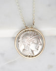 A silver pendant necklace featuring an ancient coin-style profile of a woman's face, set within a circular frame and hanging from a delicate silver chain on a light marble background.
