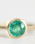 A gold ring featuring a round, faceted green emerald-cut gemstone in a prong setting, shown against a soft, light gray background.
