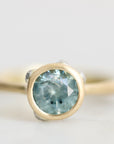 A close-up shot of a gold ring featuring a round-cut light blue gemstone in a bezel setting, with small clear accent stones on the shoulders, set against a soft, light gray background.