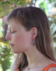 Woman with long brown hair wearing hoop earrings, standing outdoors with greenery in the background