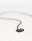 A black pendant necklace with a dark, polished rectangular stone set in a gold-toned bezel, suspended from a thin black cord against a bright white background.