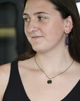 A woman wearing a dark blue teardrop-shaped drop earring and a dark necklace with a small dark pendant, shown in a close-up portrait.