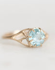 A yellow gold ring featuring a round-cut light blue aquamarine gemstone in a prong setting, accented by small clear stones along the band, shown on a plain light gray background.