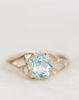 A gold ring featuring a large, round-cut light blue gemstone in a prong setting, shown on a soft, light gray background.