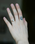 A close-up of a person's hand wearing a gold ring with a clear round gemstone on the ring finger, with a small blue geometric tattoo on the middle finger, against a dark blurred background.