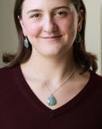 A woman wearing a silver pendant necklace with a green teardrop-shaped stone and matching green teardrop earrings, paired with a burgundy V-neck sweater.