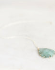 A delicate silver chain necklace featuring a leaf-shaped pendant made of textured green stone, held in a gold bezel setting, resting on a light marble surface.
