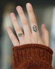 A close-up of a person's hand wearing a gold ring with a green gemstone, styled with a dark geometric tattoo on the middle finger and a rust-colored knit sweater cuff.