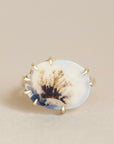 An oval ring featuring a natural agate or jasper inlay with a tree-bark pattern, held in a gold prong setting against a neutral beige background.
