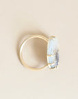 A gold ring with a round bezel-set light blue stone, shown from an angled side view on a solid light beige background.
