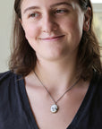 A woman wearing a silver chain necklace with a round white pendant engraved with a small black face, shown in a close-up portrait.