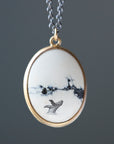 An oval pendant necklace featuring a white stone with black veining and a small whale tail illustration, set in a gold-tone bezel with a silver-tone chain.
