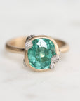 A close-up shot of a ring featuring a cushion-cut green gemstone in a gold bezel setting, accented with small white diamond-like stones on the shoulders, set against a light gray background.
