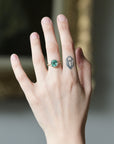 A close-up of a person's hand wearing a gold ring with a round green gemstone, set against a softly blurred indoor background.