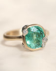 A cushion-cut green gemstone ring set in a gold-toned band with small diamond accents on the sides, photographed on a soft cream background.
