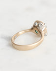 A vintage-style ring with a gold band and a silver-toned decorative top featuring a central round diamond, smaller side stones, and a tiny green accent, photographed on a light marble surface.