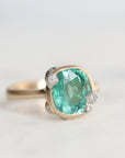 A cushion-cut green gemstone ring set in a gold-toned band with small clear accent stones, photographed on a light neutral background.
