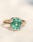 A vintage-style ring featuring a large cushion-cut green gemstone in a gold-toned setting with small clear accent stones, photographed on a light beige surface.