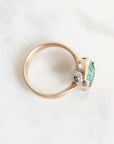 A close-up shot of a vintage-style ring with a rose gold band, featuring a green gemstone and small silver floral accents, resting on a white marble surface.
