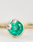 A gold ring featuring a round green emerald-cut center stone and two small silver floral accents, photographed against a soft white background.