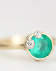A gold ring featuring a round green emerald-cut gemstone and two small clear diamond accents on the top, shown against a soft white background.