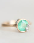 A close-up shot of a ring featuring a round, faceted light green gemstone in a gold-toned bezel setting, accented with small platinum barnacles on the sides, set against a soft, light gray background.