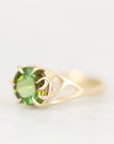 A yellow gold ring featuring a round green peridot gemstone in a prong setting, with a small clear accent stone on the band, shown against a plain white background.