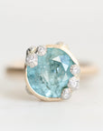 A close-up shot of a ring featuring a large, faceted light blue gemstone in a gold-toned setting, accented by several small round clear stones, set against a plain light background.
