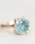 A close-up shot of a ring featuring a large oval light blue gemstone in a gold organic bezel accented with small accent barnacles, on a light gray background.