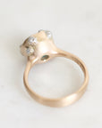 A close-up shot of a gold ring with a textured, organic-shaped setting featuring three small clear gemstones, resting on a light marble surface.