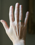 A close-up of a person's hand wearing a gold ring with a round green gemstone, set against a softly blurred indoor background.