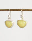 A pair of silver dangle earrings with smooth, polished light yellow-green stone pendants, hanging from a horizontal brass rod against a plain white background.